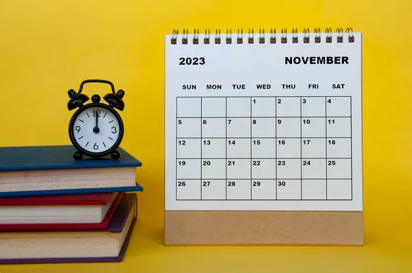 November 2023 desk calendar with books and alarm clock on yellow background. Calendar concept