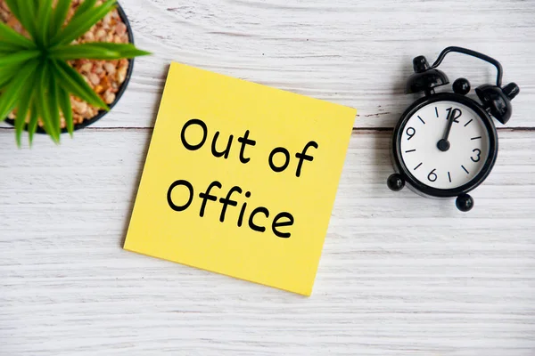 Out of office text on sticky note with alarm clock and plant background. Business concept