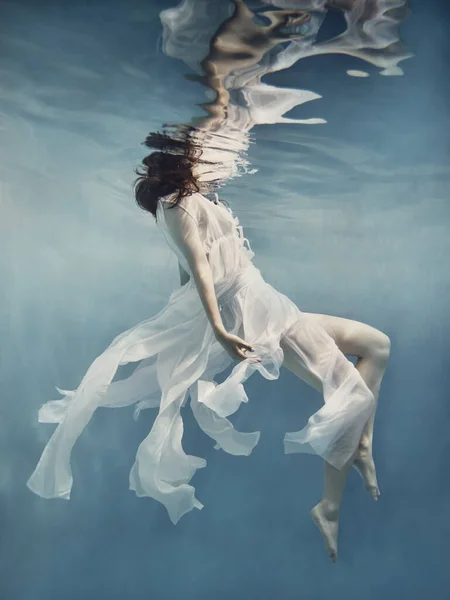 白いドレスを着た暗い髪の女の子がまるで無重力の中を飛んでいるかのように水中で泳ぐ — ストック写真