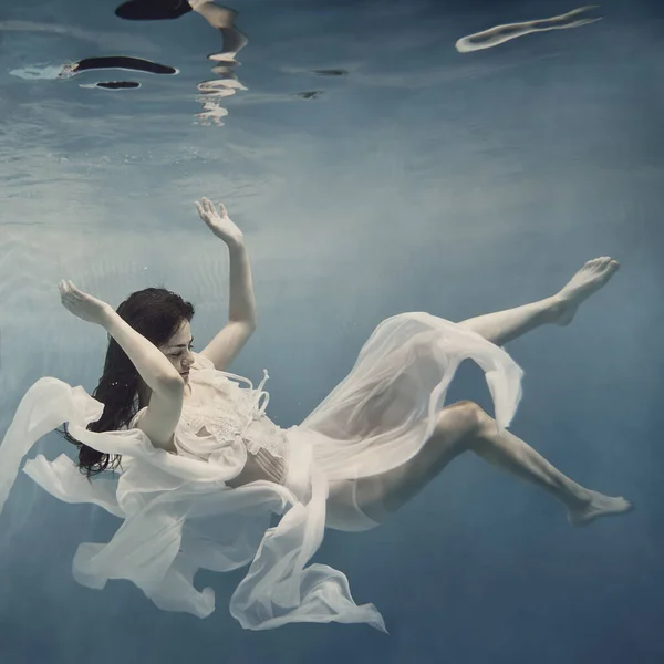 白いドレスを着た暗い髪の女の子がまるで無重力の中を飛んでいるかのように水中で泳ぐ — ストック写真