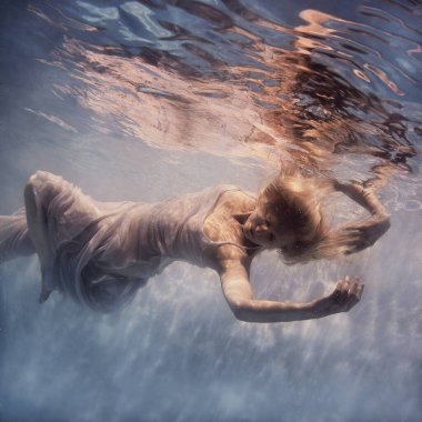      Beyaz elbiseli, sarı saçlı bir kadın sanki yer çekimsiz havada yüzüyormuş gibi suyun altında yüzüyor.                          