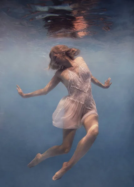 白いドレスを着た金髪の女がまるで無重力の中を飛んでいるかのように水中で泳ぐ — ストック写真