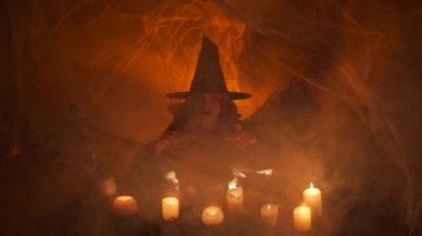A teenager disguised as a witch casts a spell over a ritual cauldron, she is enveloped in thick smoke. Halloween concept. Scary story, bedtime stories. Witch's house