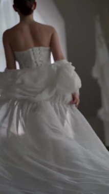 Dikey videolar. Güzel bir gelin uzun bir trenle beyaz bir bornozla pencereye doğru gidiyor, seksi görünüyor.