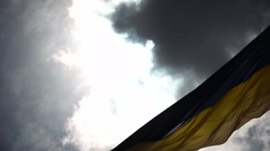 The blue-yellow flag with the national colors of Ukraine flutters against the background of dark clouds. The sun hides behind a dark cloud. Looks epic.