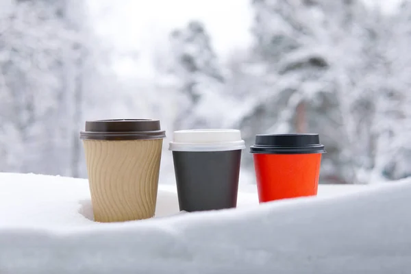 Ormanın arka planında kar olan üç bardak kahve. Kırmızı siyah ve bej kağıt bardaklar. Boşluğu kopyala