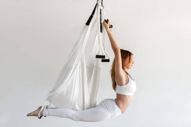 Sinek yogası pratiği yapan genç bir kadın. Spor salonundaki gri arkaplanı esnetiyorum. Spor ve sağlıklı yaşam tarzı kavramı. Beyaz spor giyim.