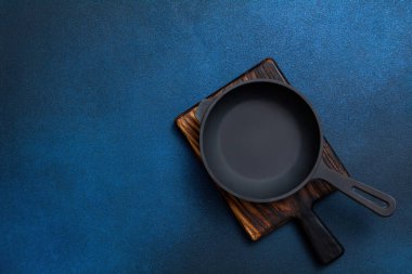 Cast iron skillet on a wooden board. Frying pan and cutting board on a dark blue background. Kitchen utensils. Empty place. Top view.