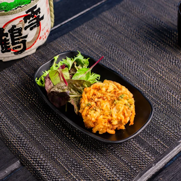 Veggie vegan tempura with salad in the traditional Japanese ramen restaurant, with a black plate on a black mat and Japanese fonts sake barrel translation: Snow Crane