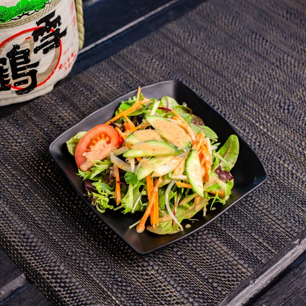 Japanese salad with tomato, cucumber and sesame dressing in the traditional Japanese ramen restaurant, with a black plate on a black mat and Japanese fonts sake barrel translation: Snow Crane