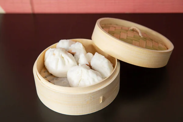 Char siew bao steamed buns with BBQ pork in a bamboo steam basket container of traditional Cantonese yum-cha Asian gourmet cuisine meal food dish on the white serving plate and brown red table