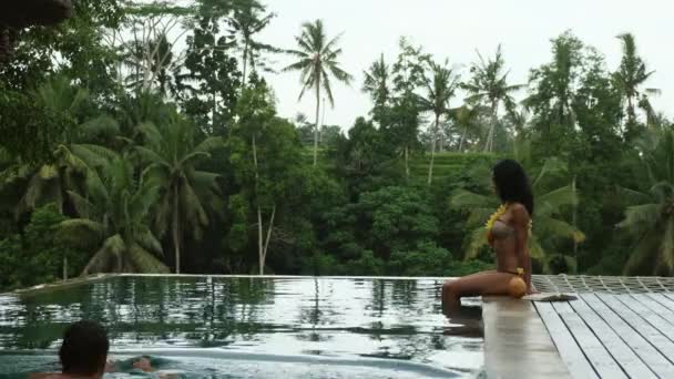 Egy karcsú lány bikiniben ül a medence mellett. A fickó úszik. dzsungel Bali — Stock videók