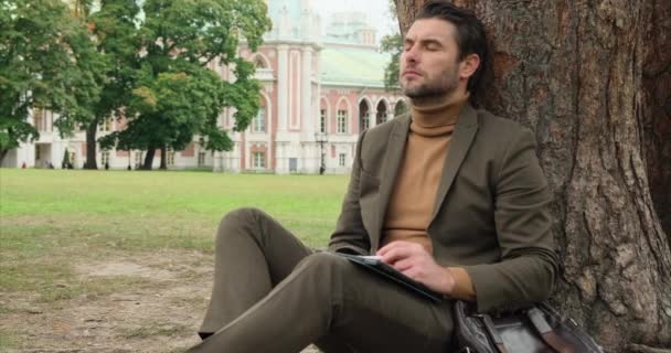 Hombre de negocios guapo usando tableta sentada cerca de un árbol en el parque bingo de Viena — Vídeos de Stock