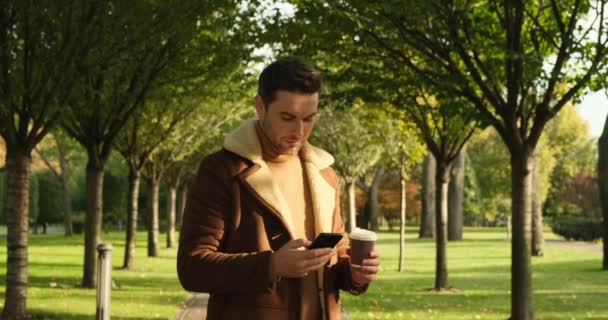 Usmívající se atraktivní muž v Braunově kabátě s telefonem jít do podzimního parku — Stock video