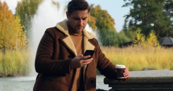Ελκυστικός άντρας με παλτό με καφέ πίνει καφέ κοντά στη λίμνη και σιντριβάνια — Αρχείο Βίντεο