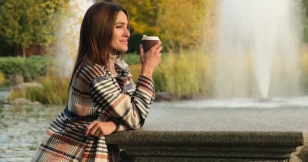 Attractive woman in coat with Coffee drinks coffee near the lake and fountains — Stock Video