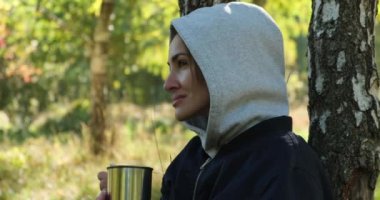 Ormanda bir kadın kütüğün üzerinde oturup sıcak bir şeyler içiyor. Bir sepet mantar.
