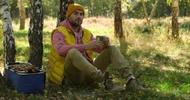 Чоловік у лісі сидить на пні, п'є гарячий напій. кошик з грибів — стокове відео