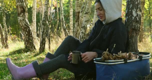 Une femme dans la forêt est assise sur un moignon en train de boire une boisson chaude. panier de champignons — Video