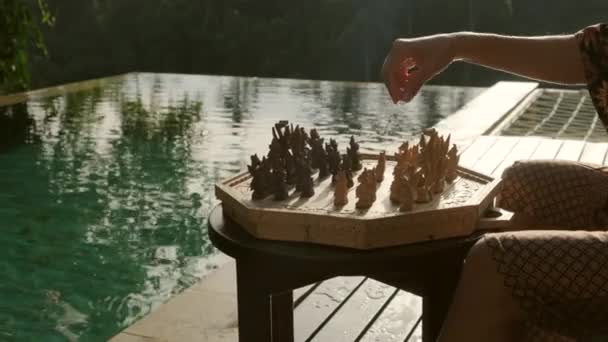 Chica en un albornoz juega ajedrez hecho a mano cerca de la piscina en una villa tropical — Vídeos de Stock