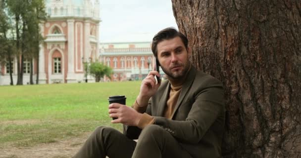 Человек во время обеда в городском парке сидит с кофе и разговаривает по телефону — стоковое видео