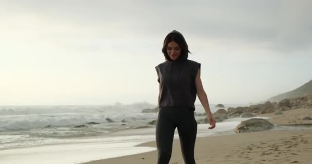 Menina esportiva em um capuz caminha ao longo da praia perto do oceano após o treinamento — Vídeo de Stock