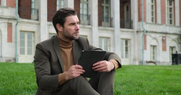 Om de afaceri într-un costum stă într-un parc lângă un copac cu o tabletă și scrie un raport — Videoclip de stoc