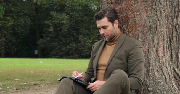 Empresario en un traje se sienta en un parque cerca de un árbol con una tableta y escribe informe — Vídeos de Stock