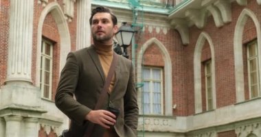 Omuz çantalı şık bir adam kahve içiyor ve başka tarafa bakıyor.