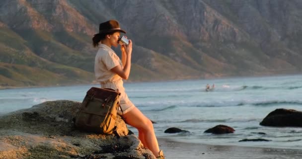 Menina senta-se nas montanhas da costa do oceano e bebe uma bebida quente de uma caneca de ferro — Vídeo de Stock