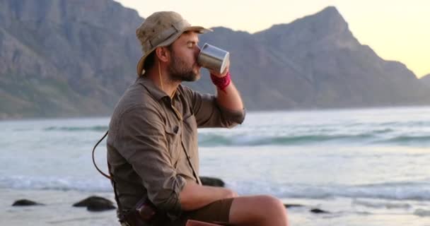 O homem senta-se nas montanhas da costa do oceano e bebe uma bebida quente de uma caneca de ferro — Vídeo de Stock