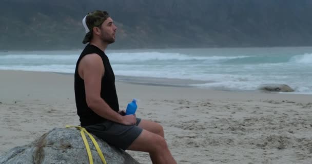 Atlet pije vodu po tréninku, zatímco sedí na kameni ne na pláži oceánu — Stock video