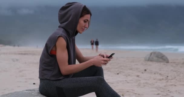 Kaukasische athletische junge Frau lächelt mit ihrem Smartphone in der Nähe der Ozeanwelle — Stockvideo
