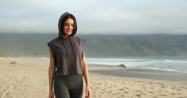 Chica deportiva en una campana camina a lo largo de la playa de la mañana cerca de las montañas del mar — Vídeo de stock