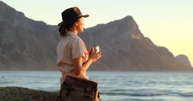 Kız okyanus kıyısındaki dağlarda oturur ve demir bardaktan sıcak bir içecek içer.