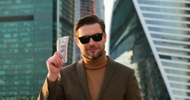 Чоловік у костюмі та сонцезахисних окулярах показує долари камері. молодий мільйонер — стокове відео