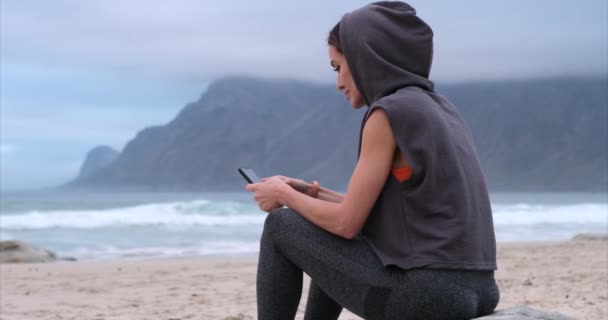 Caucasien athlétique jeune femme souriant en utilisant son téléphone intelligent près de Ocean wave — Video