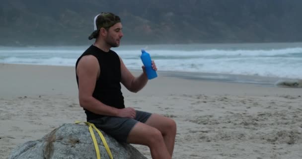 Deportista hombre beber agua después de entrenar sentado en una piedra no en la playa — Vídeo de stock
