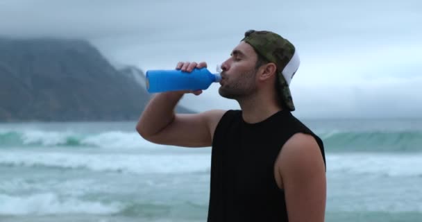 Atleta muscoloso beve acqua da una bottiglia sulla riva dell'oceano dopo una tempesta — Video Stock