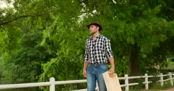 有料のシャツを着た男が田舎の農場で空の箱を持って歩いている — ストック動画