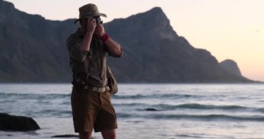 Sırt çantalı turist, Afrika safarisi tarzında, okyanus kıyısında fotoğraf çekiyor.