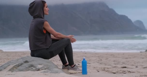 Chica atlética en ropa deportiva se sienta en una roca en la playa, disfruta de la puesta de sol — Vídeo de stock