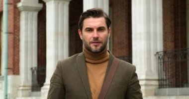 Omzunda çanta olan kahverengi takım elbiseli bir adam kameraya bakıyor.