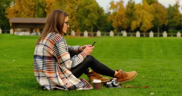 Meisje zit op groen gras in het park met telefoon en koffie — Stockvideo