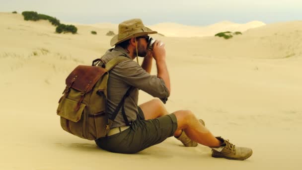 A man in safari clothes sits on the sand in the desert and drinks the of water — Vídeo de stock