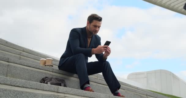 Man in a business suit sits on the steps near the office center with a phone — Video Stock