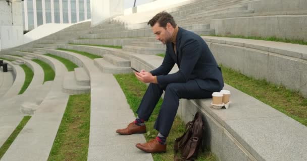 Man in a business suit sits on the steps near the office center with a phone — Vídeo de stock