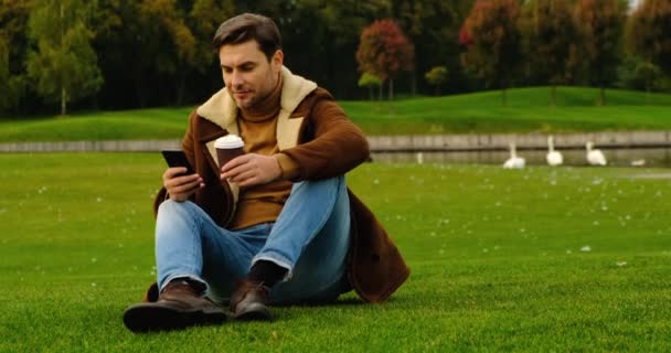 身穿夹克和牛仔裤的英俊男子坐在公园的草地上喝咖啡 — 图库视频影像