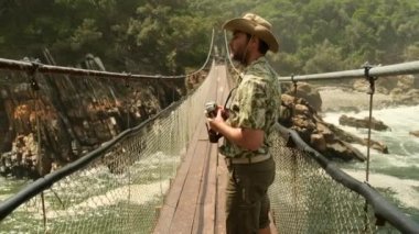 Dağlar arasında asılı bir köprüden geçen bir adam ipi. Tsitsikama Parkı