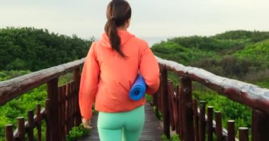 Turuncu rüzgarlıklı atletik bir kız, okyanus kıyısında yoga minderiyle ahşap bir köprü boyunca yürüyor. Sportif formda genç bir kadın spor giysisi giyer. Elinde yoga sarılı lastik minder taşır.